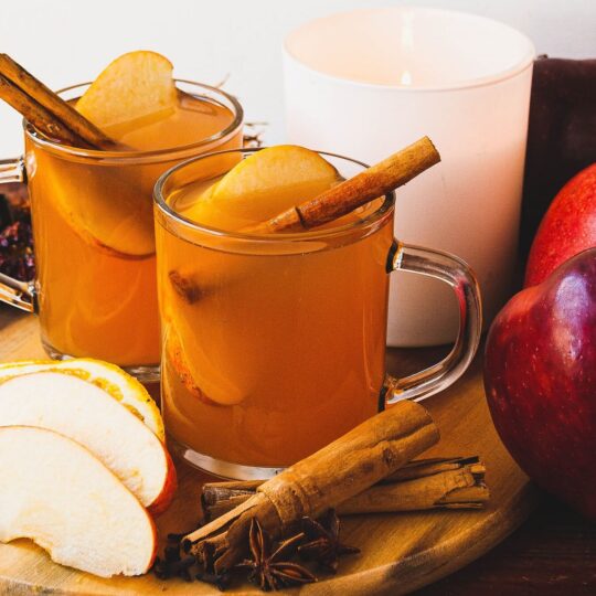Two glass mugs of hot apple cider garnished with cinnamon sticks and apple slices.