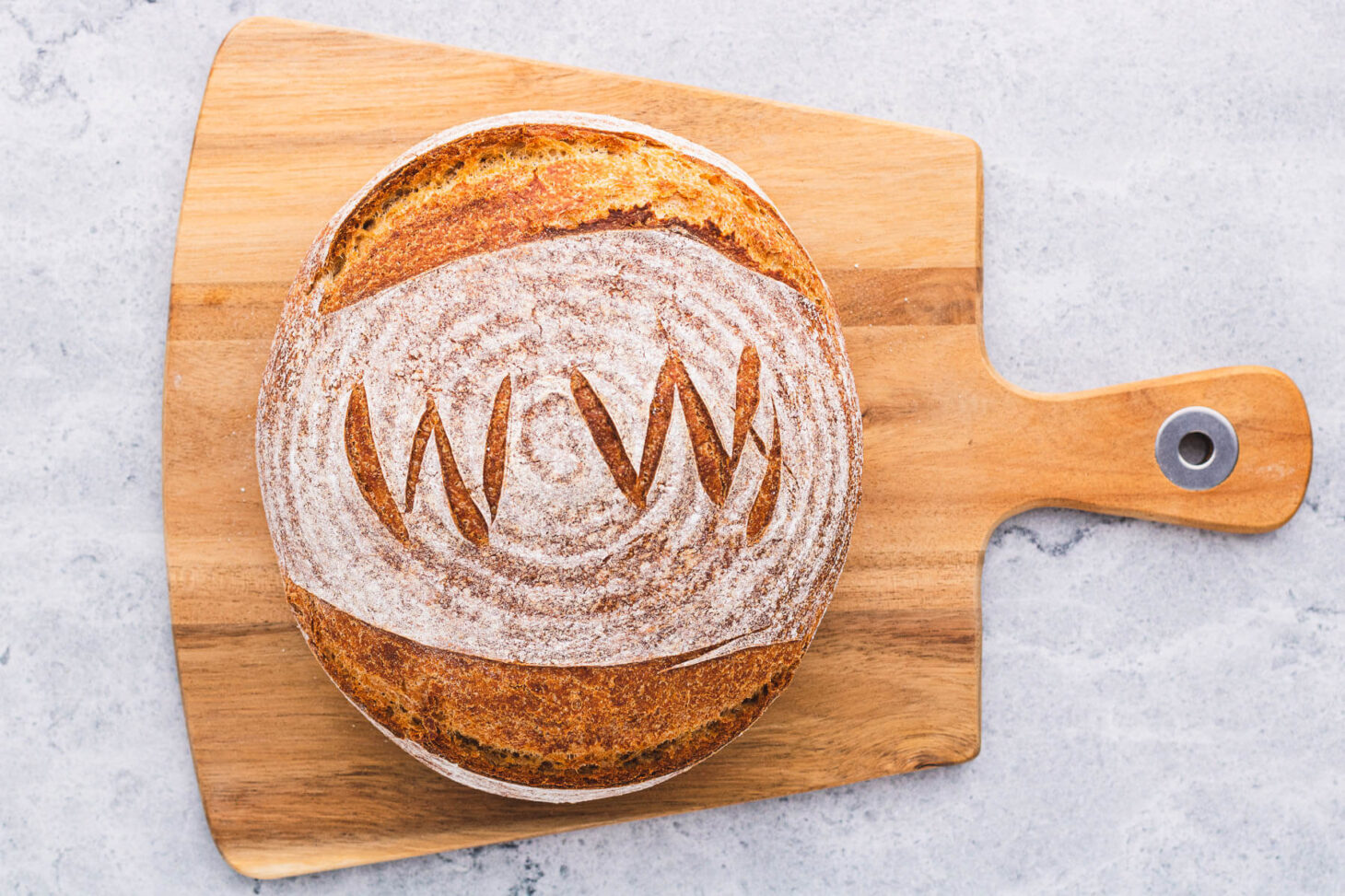 A boule of No Knead Whole Wheat Bread marked with WW scoring pattern on a wooden cutting board.