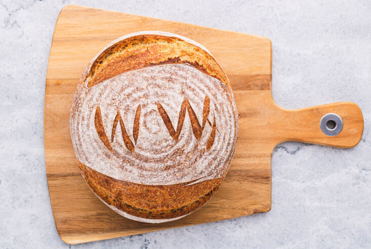 A boule of No Knead Whole Wheat Bread marked with WW scoring pattern on a wooden cutting board.