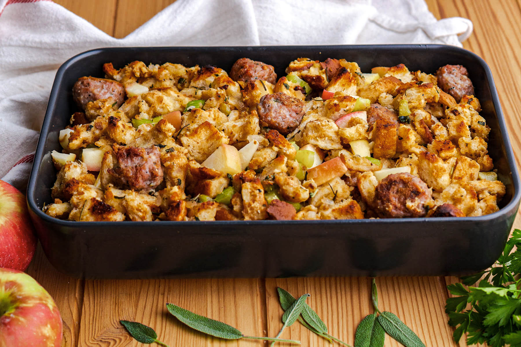 A grey baking dish full of golden baked Apple Sausage Stuffing beside fresh Italian Parsley, sage, and red apples.