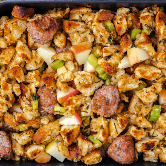 A grey baking dish full of golden baked Apple Sausage Stuffing beside Italian Parsley and red apples.