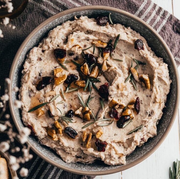 A grey bowl filled with white bean hummus garnished with walnuts, dried cranberries, and fresh rosemary.