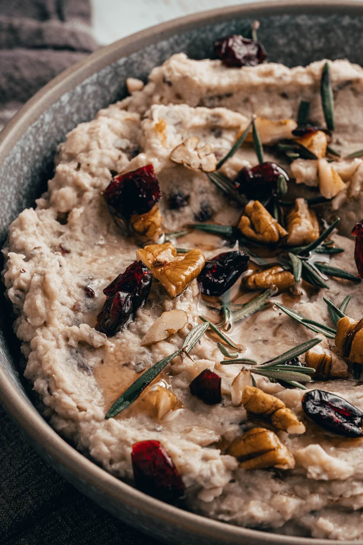 A grey bowl filled with white bean hummus garnished with walnuts, dried cranberries, and fresh rosemary.