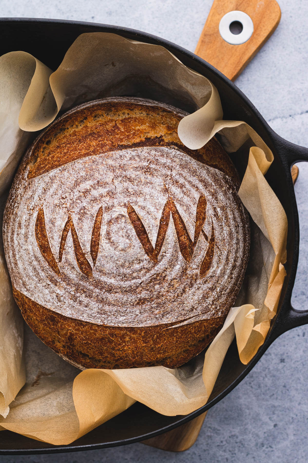 A boule of No Knead Whole Wheat Bread marked with WW scoring pattern in a bed of parchment paper in a black Dutch oven.