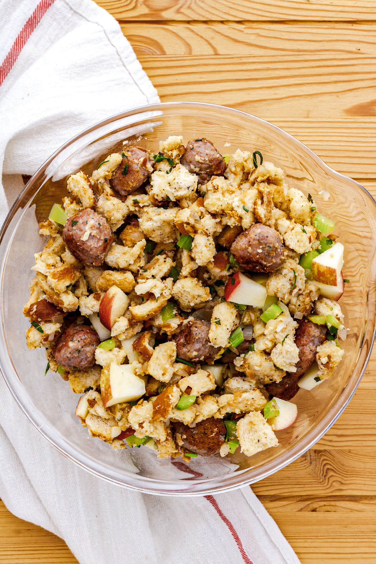A glass mixing bowl full of unbaked Apple Sausage stuffing ingredients.