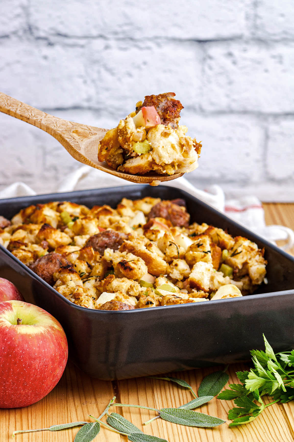 A wooden spoon full of Apple Sausage Stuffing hovers over a pan of the same stuffing.