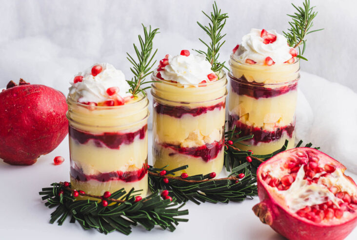 Three glass half pint jars filled with a layered Christmas trifle garnished with whipped cream, pomegranate seeds, and fresh rosemary.