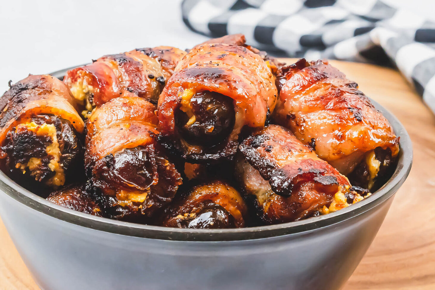 A bowl of glistening bacon wrapped stuffed dates on a wooden board.