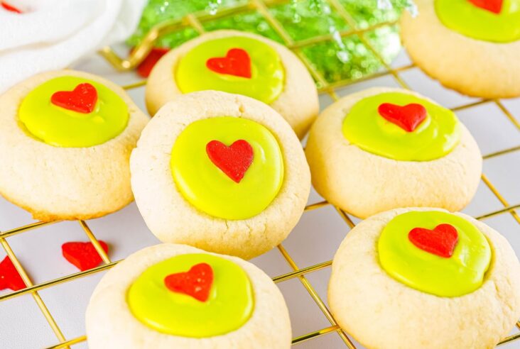 Golden baked thumbprint grinch cookies filled with bright green candy melts and decorated with a red heart on a wire cooling rack.