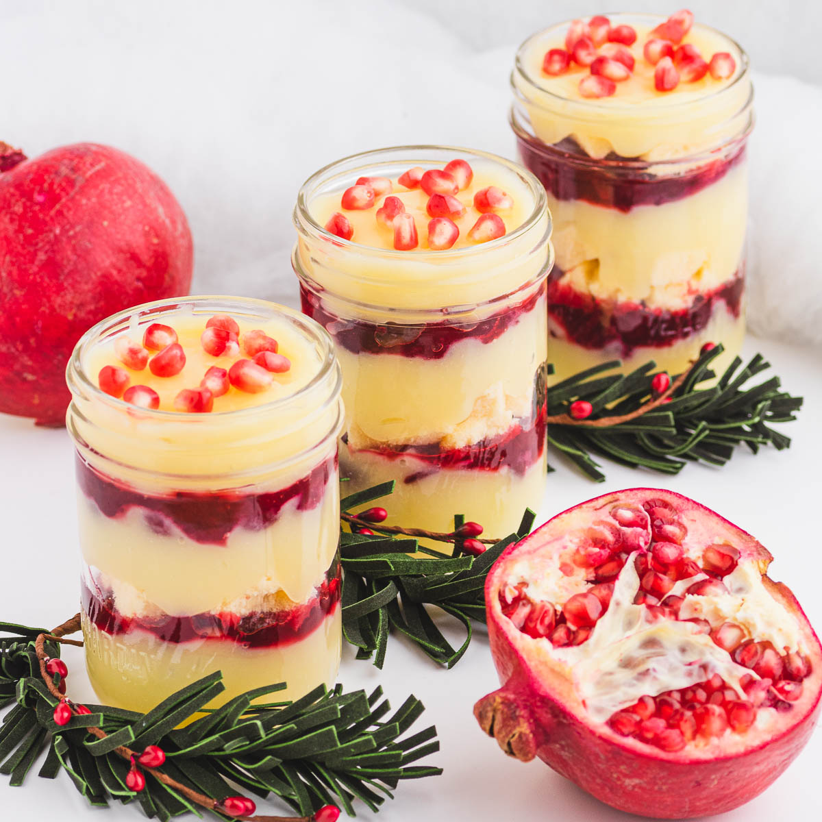 Three glass half pint jars filled with a layered Christmas trifle garnished with pomegranate seeds.