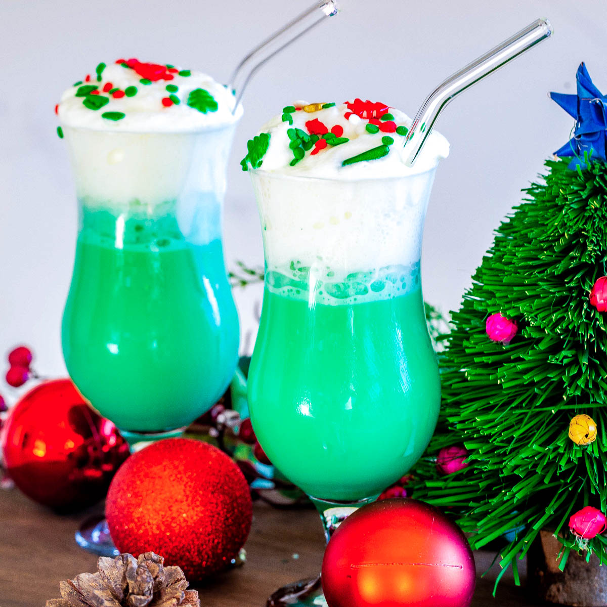 Two hurricane glasses filled with blue hawaiian cocktail topped with whipped cream and christmas sprinkles beside a tiny christmas tree.