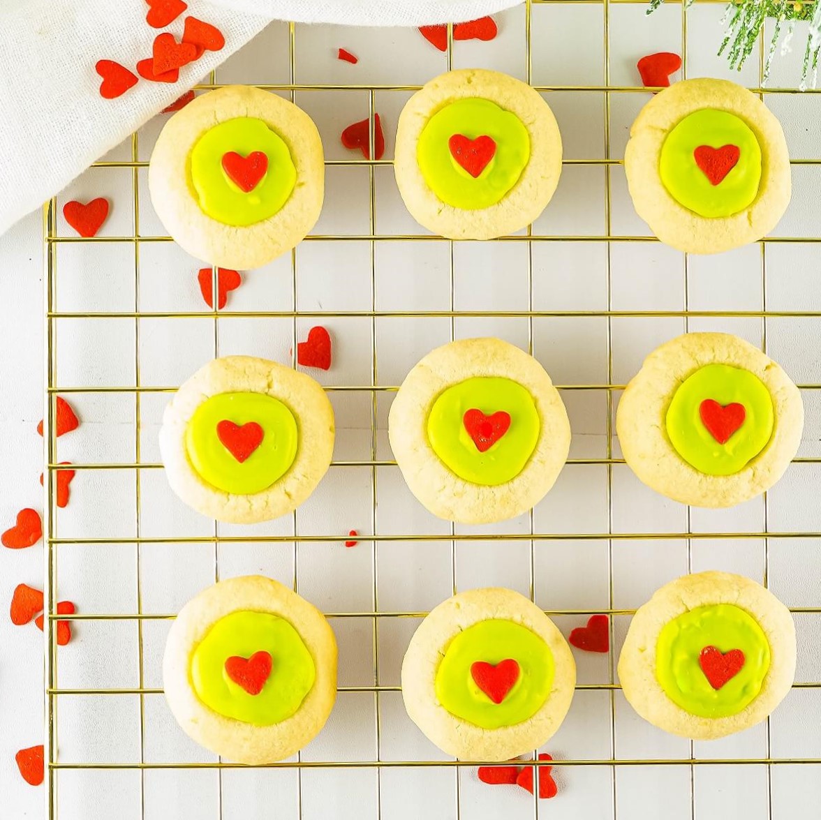 Golden baked thumbprint grinch cookies filled with bright green candy melts and decorated with a red heart on a wire cooling rack.