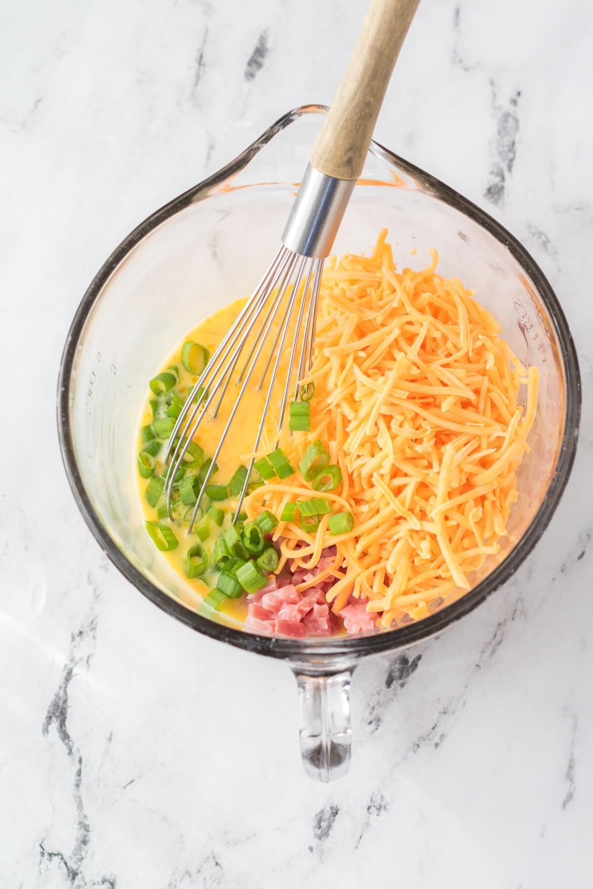 A large glass measuring cup containing whisked eggs, green onions, cheese, and ham.