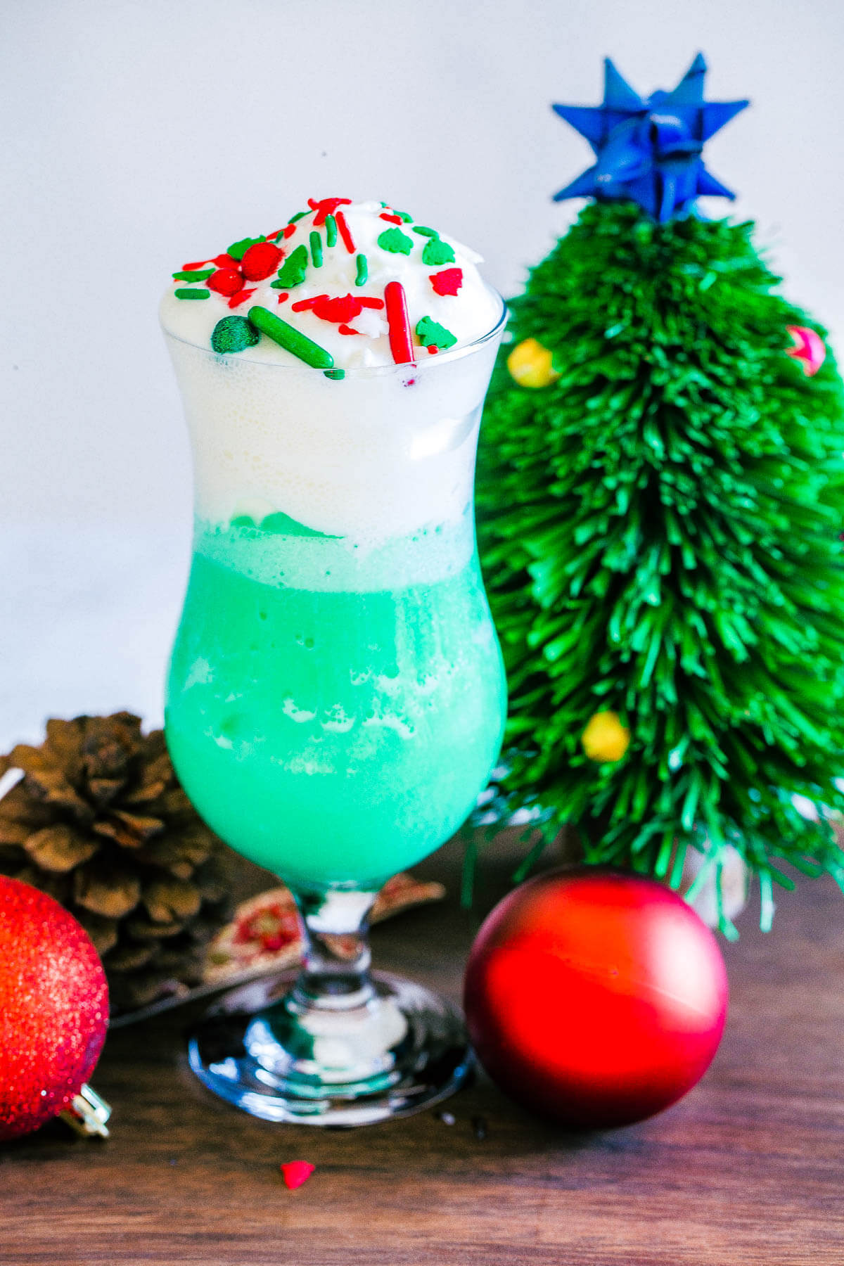 A hurricane glass filled with blue hawaiian cocktail topped with whipped cream and christmas sprinkles beside a tiny christmas tree.