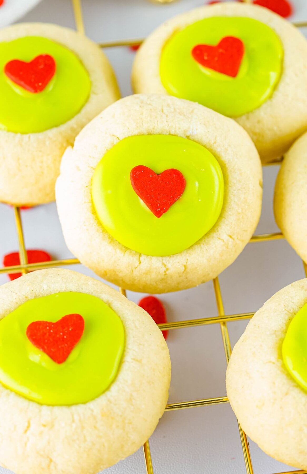 Golden baked thumbprint grinch cookies filled with bright creamy green candy melts and decorated with a red heart on a wire cooling rack.