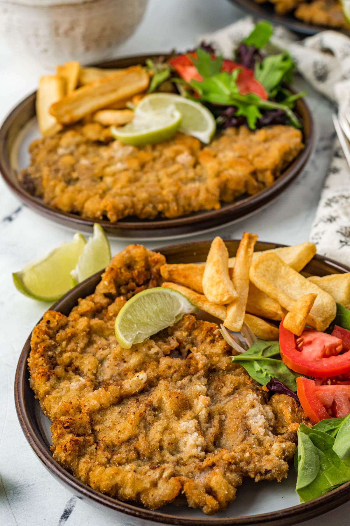 Two plates full of golden fried Milanesa de res with chips and a salad.