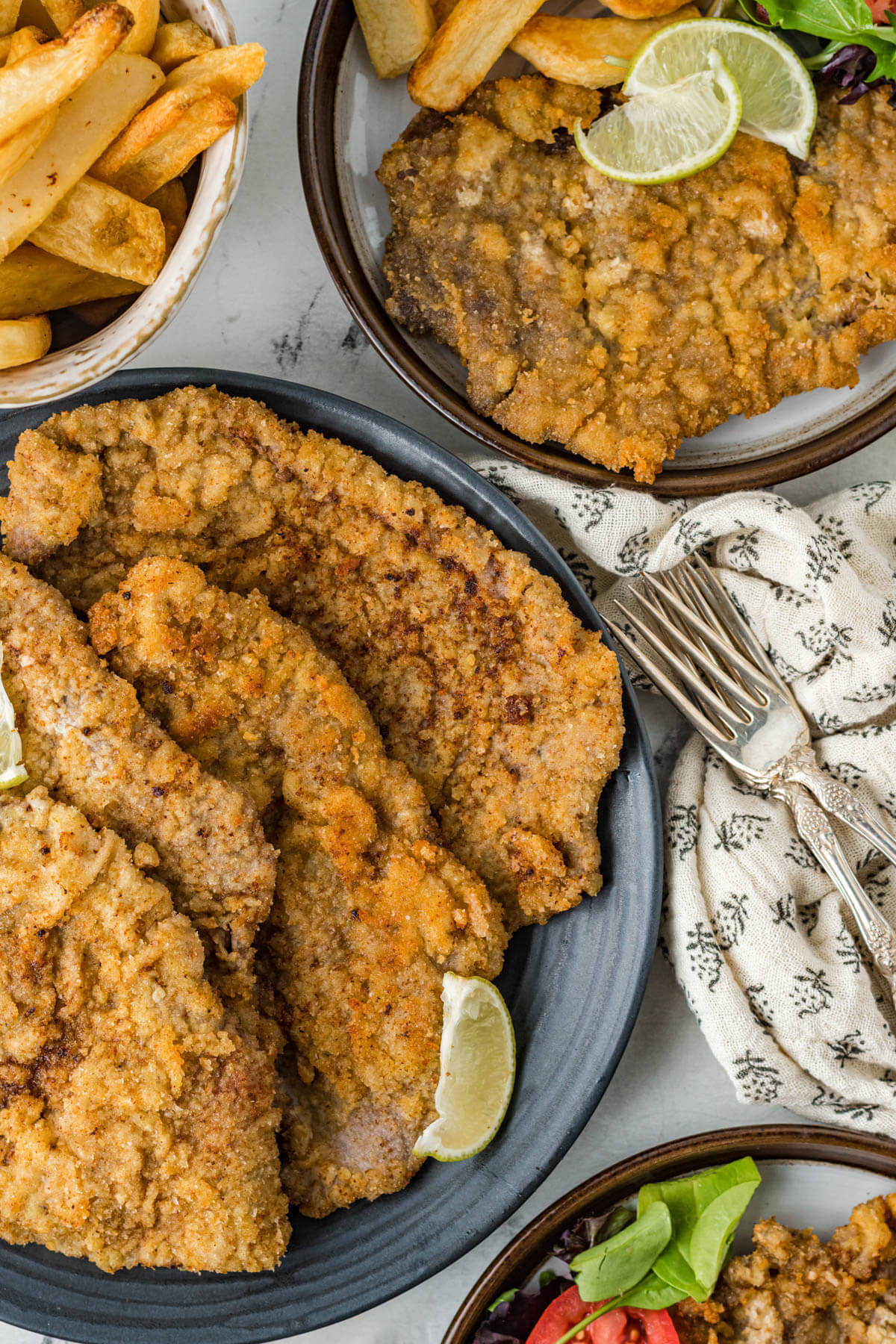 Golden fried Milanesas de Res arranged on a blue serving platter with wedges of lime.
