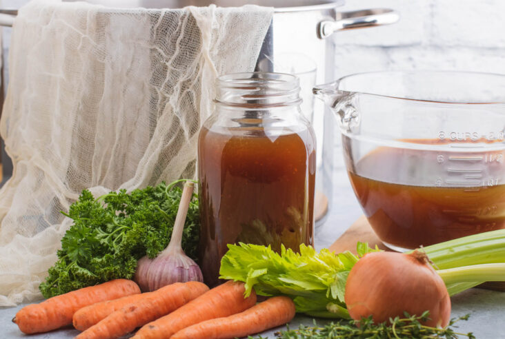 A glass quart jar filled with beef stock surrounded by fresh vegetables.
