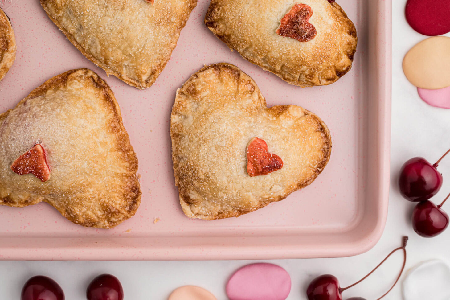 A pink baking sheet full of golden baked heart shaped individual cherry hand pies decorated with red pastry hearts.