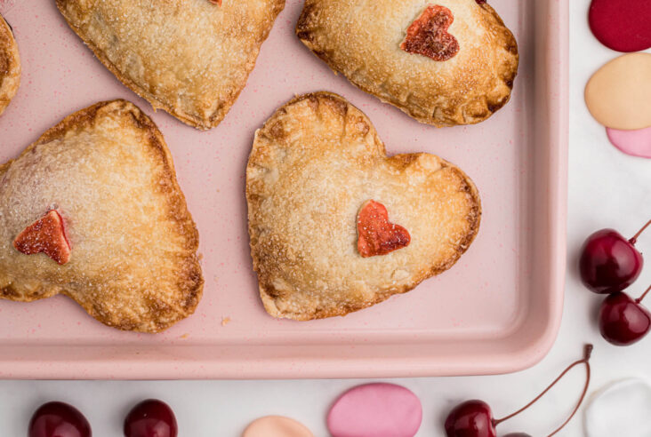 A pink baking sheet full of golden baked heart shaped individual cherry hand pies decorated with red pastry hearts.