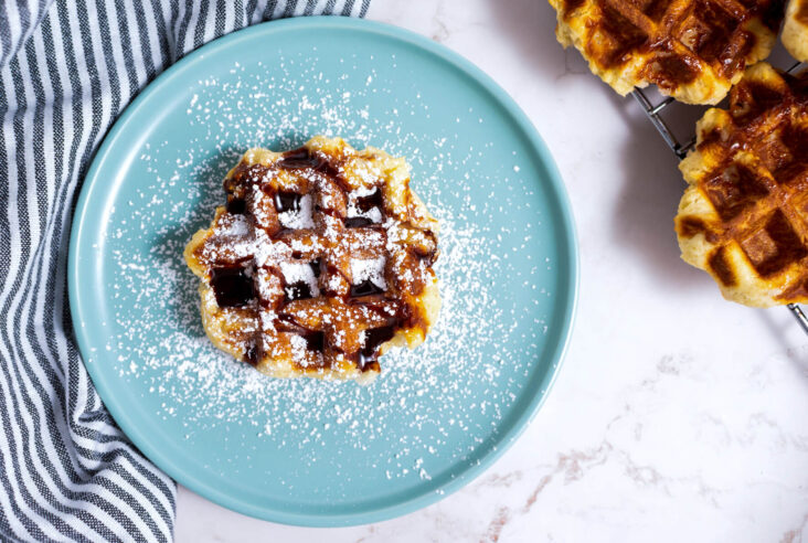 One Belgian Liège Waffle with confectioners' sugar and chocolate sauce sitting on a blue plate beside a stack of more waffles.