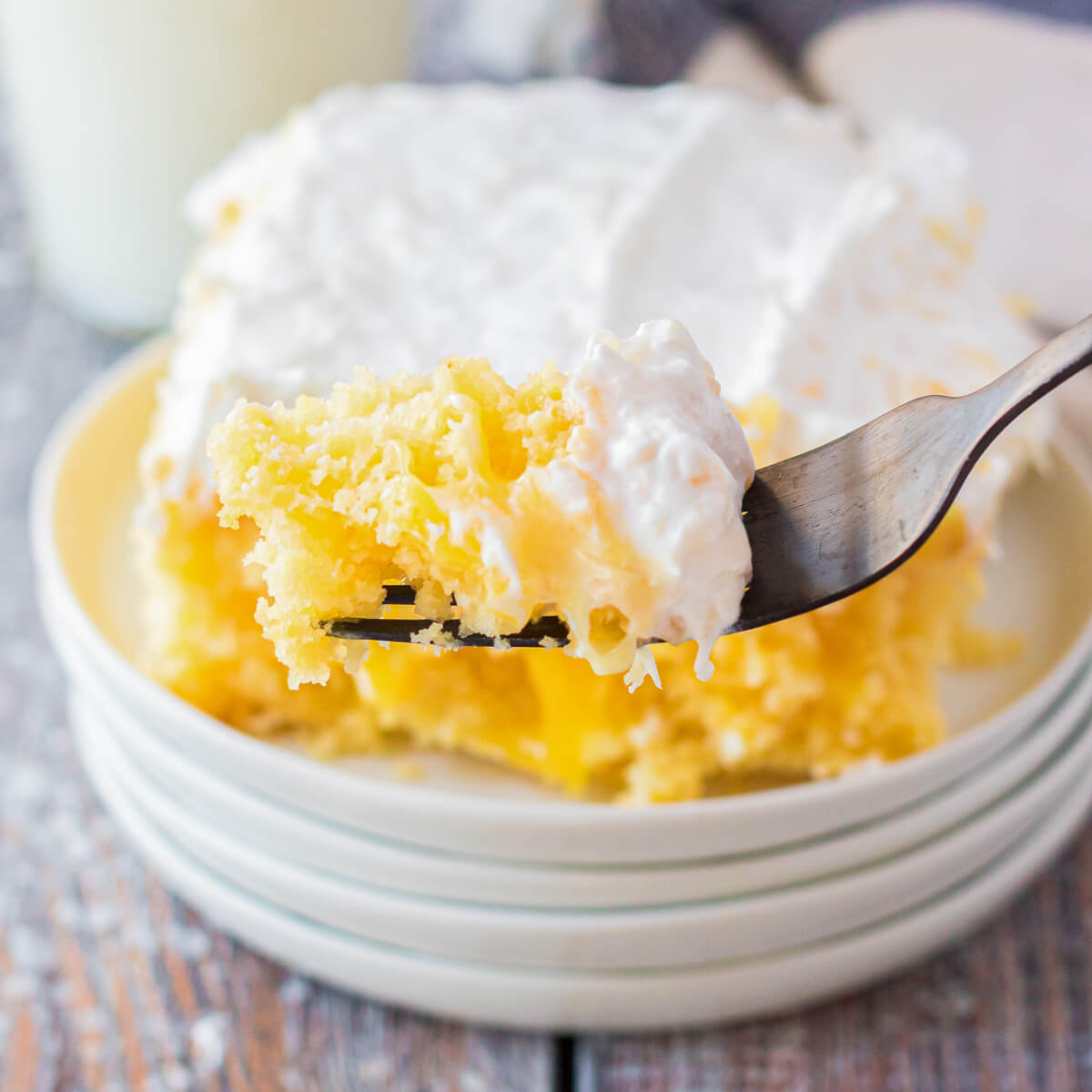 A fork holds a bite of cake in front of a square of dreamy Pineapple Poke Cake with creamy whipped topping on a stack of white plates.