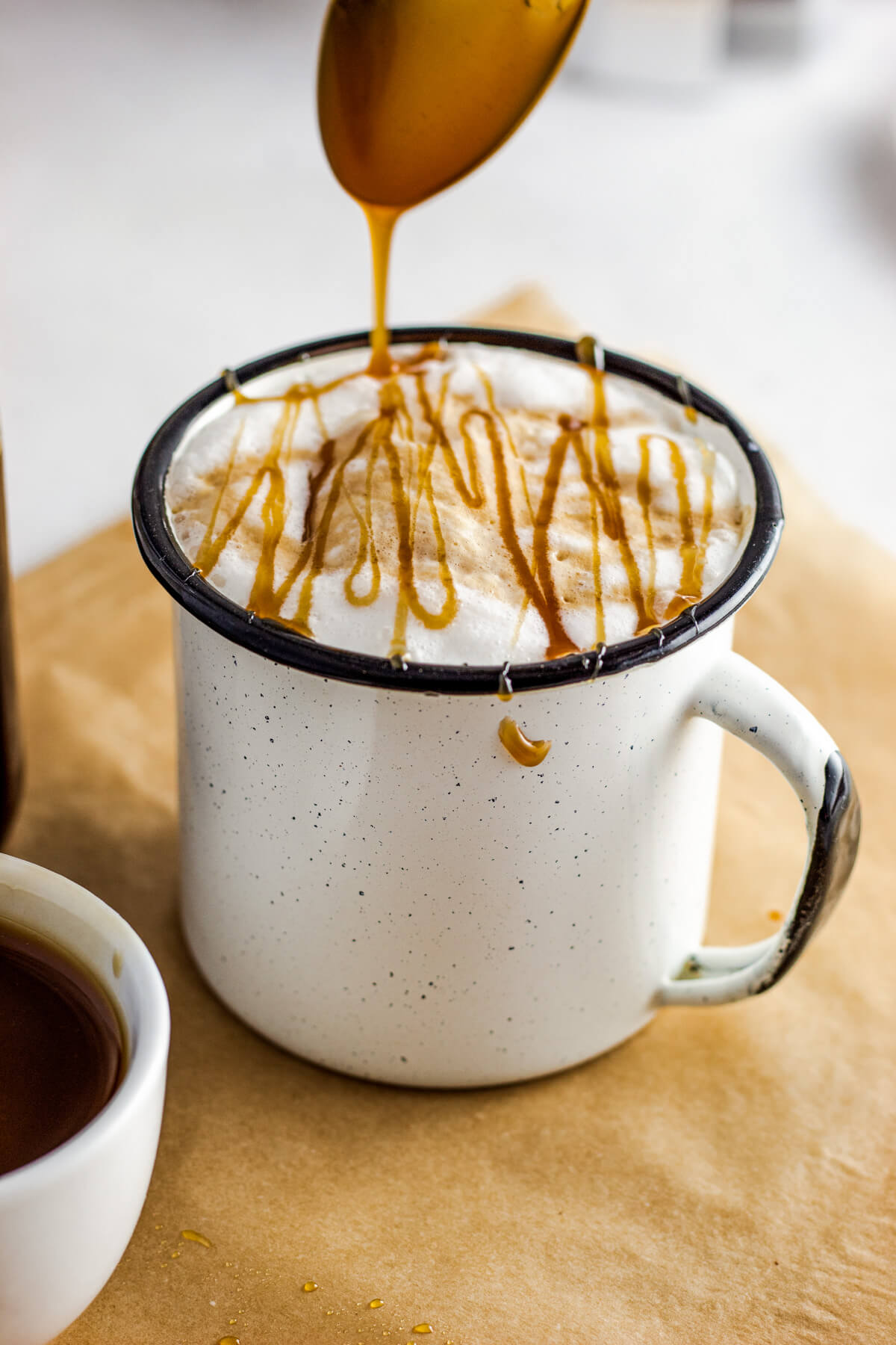 A spoon drizzles caramel sauce over a mug of coffee with foam on top.