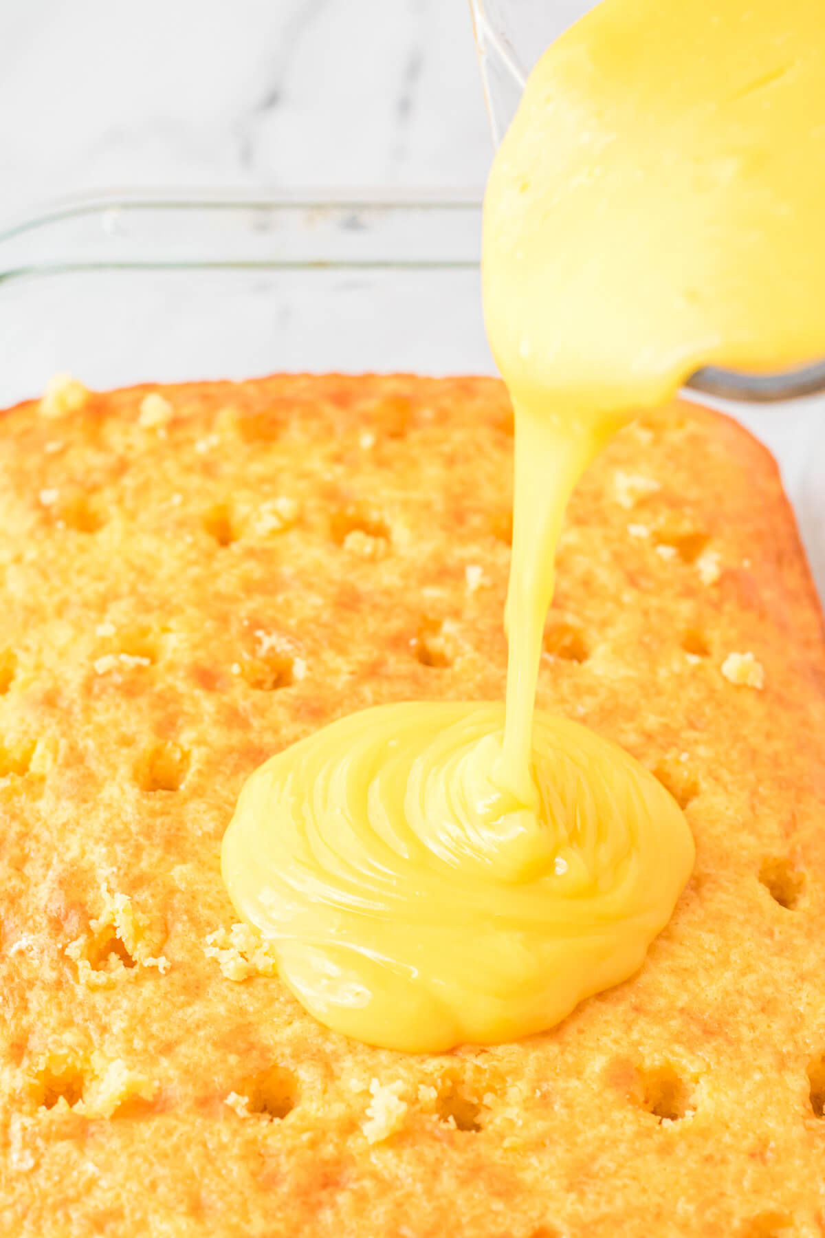Pineapple vanilla pudding being poured over a cake with holes poked in it.