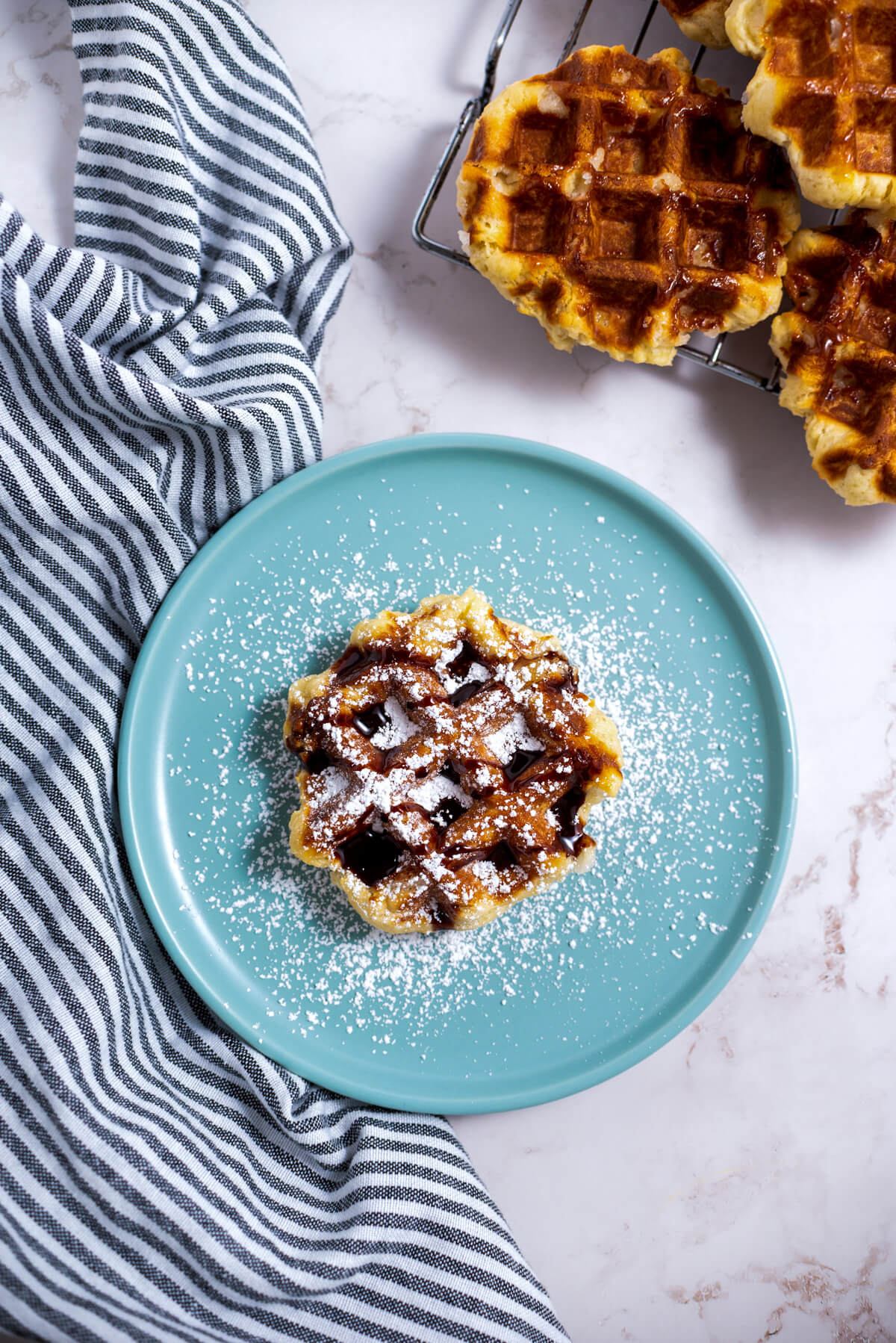 One Belgian Liège Waffle with confectioners' sugar and chocolate sauce sitting on a blue plate beside a stack of more waffles.