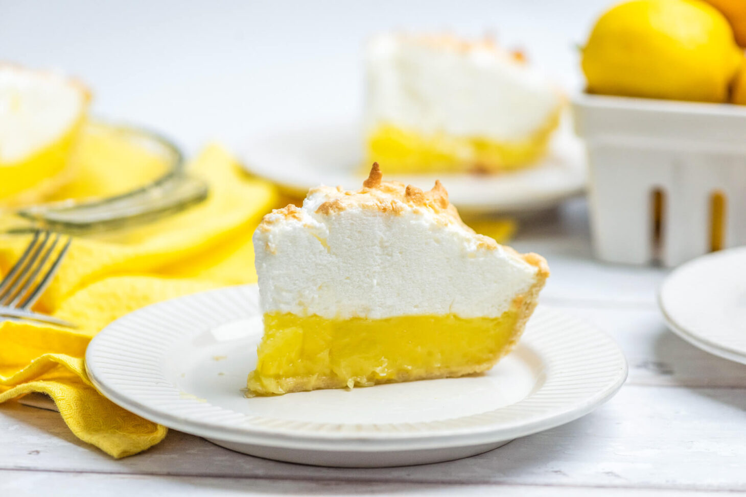 A slice of lemon meringue pie surrounded by lemons and a yellow napkin.