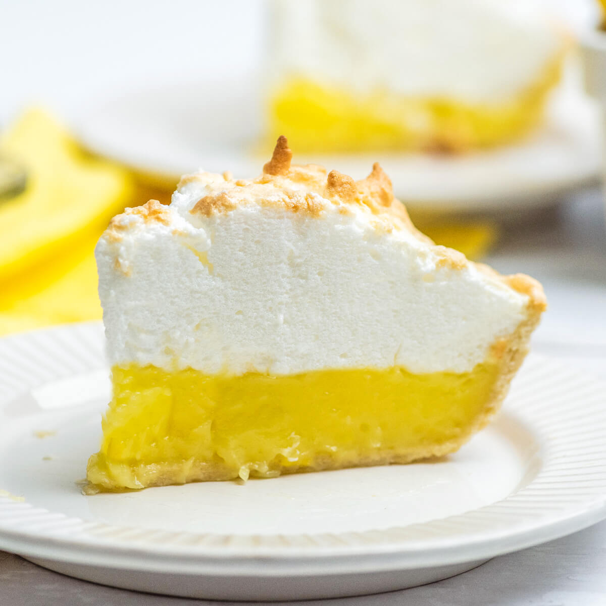 A slice of lemon meringue pie surrounded by lemons and a yellow napkin.