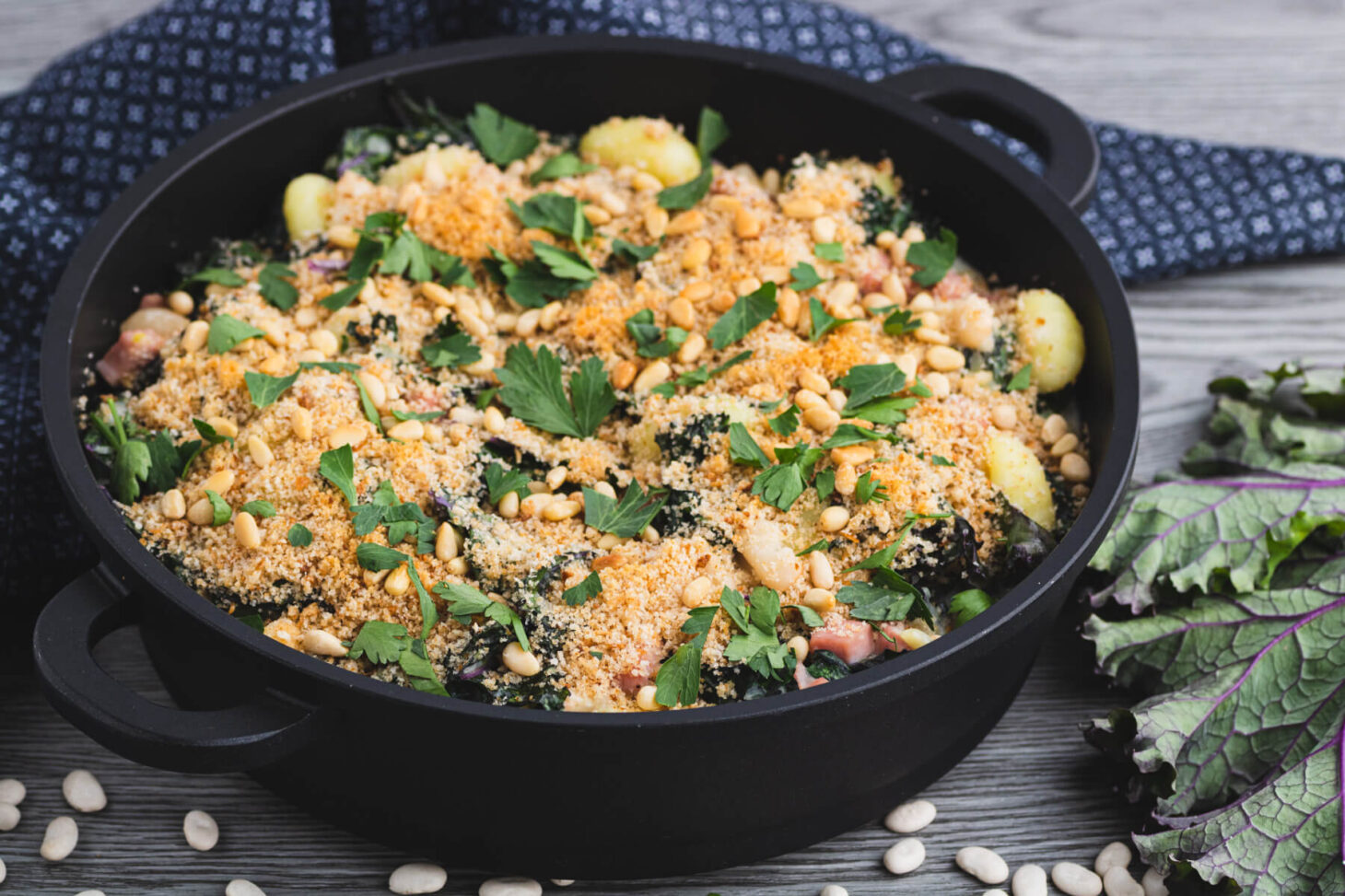 A baking dish full of Creamed Kale Gnocchi full of ham and beans in a creamy sauce topped with golden breadcrumbs and pine nuts.