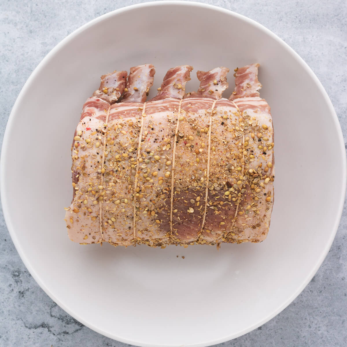 A raw pork rib roast coated with crushed coriander seeds in a white bowl.