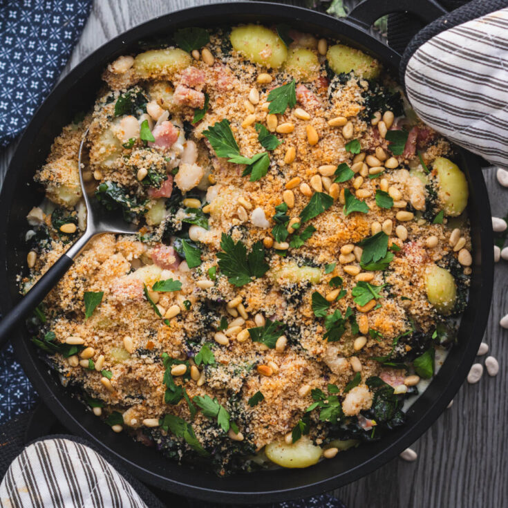 A baking dish full of Creamed Kale Gnocchi full of ham and beans in a creamy sauce topped with golden breadcrumbs and pine nuts.