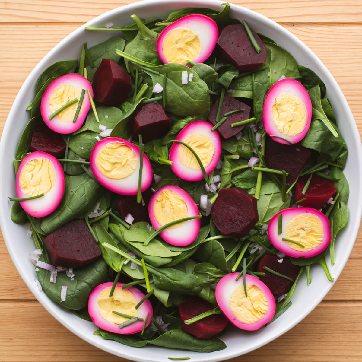 A white salad bowl filled with pickled beets, bright pink eggs and spinach salad