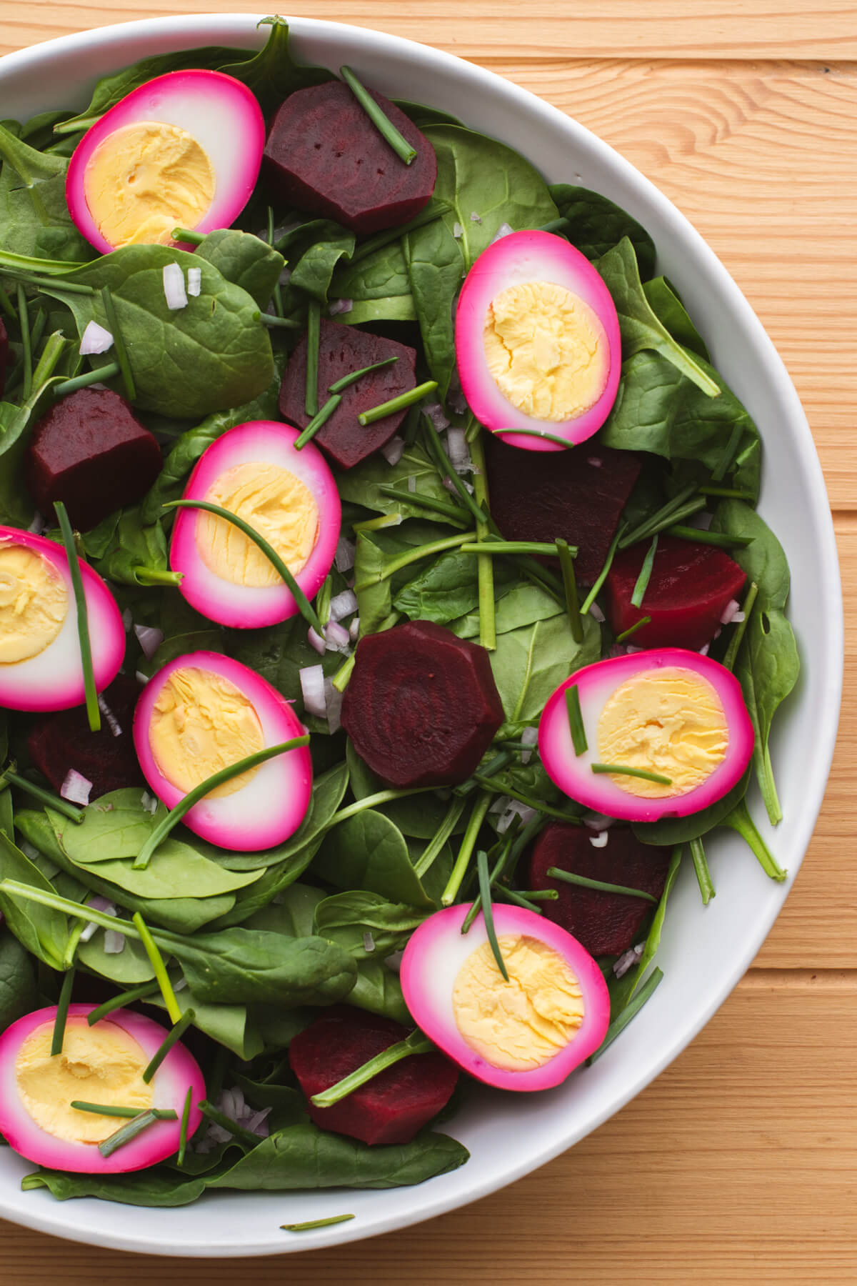 A white salad bowl filled with pickled beets, bright pink eggs and spinach salad