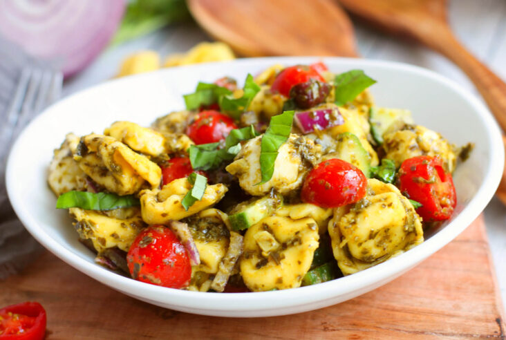 A white bowl filled with brightly coloured Pesto Tortellini Salad with bocconcini, cherry tomatoes, pesto, cucumbers, and basil.