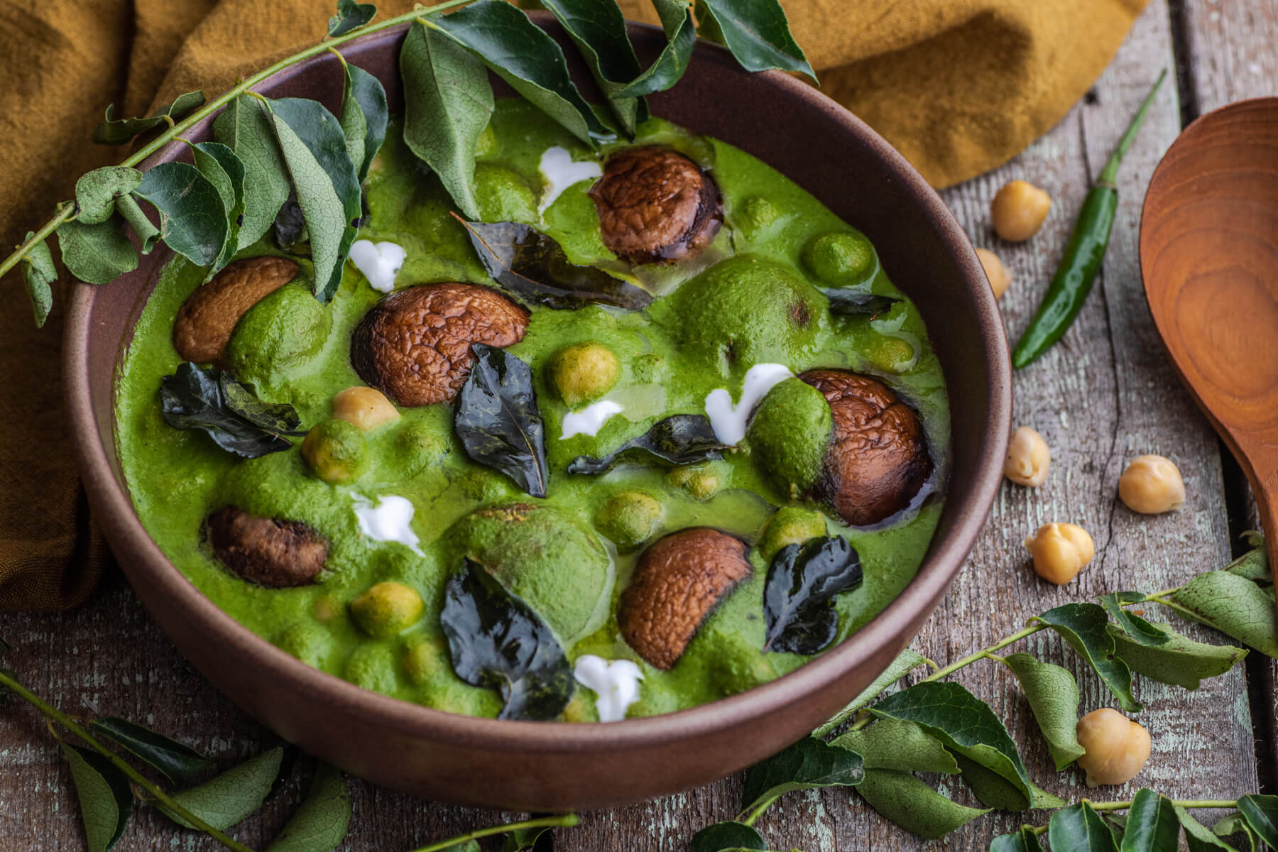 A vibrant green chana saag with mushrooms, chickpeas, fried curry leaves, and drizzles of coconut cream in a brown bowl.