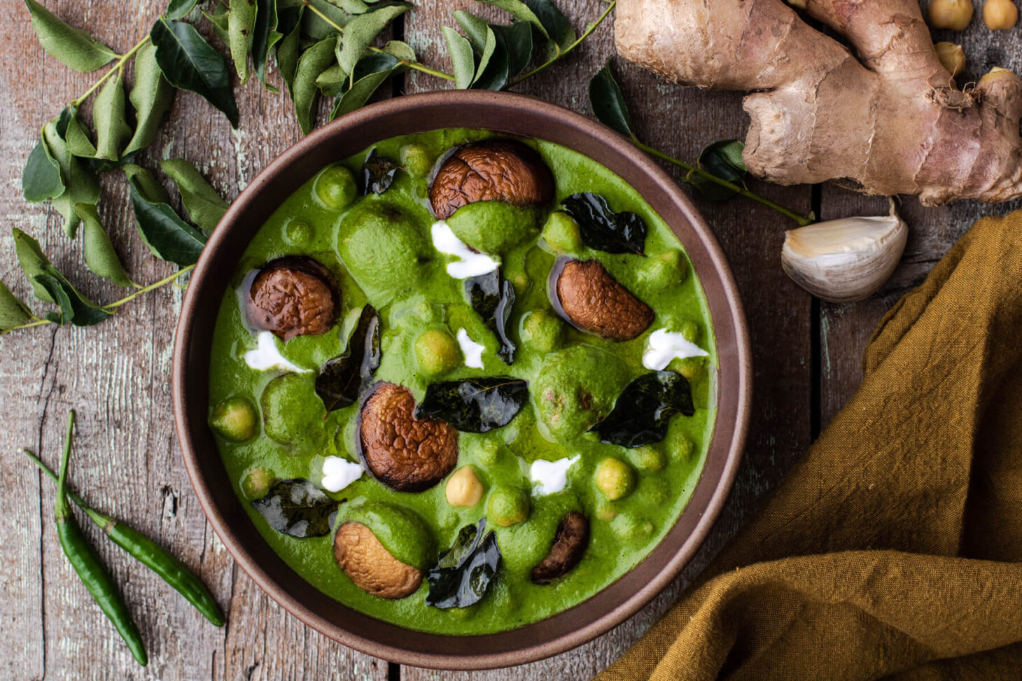 A vibrant green spinach curry with mushrooms, chickpeas, fried curry leaves, and drizzles of coconut cream in a brown bowl.