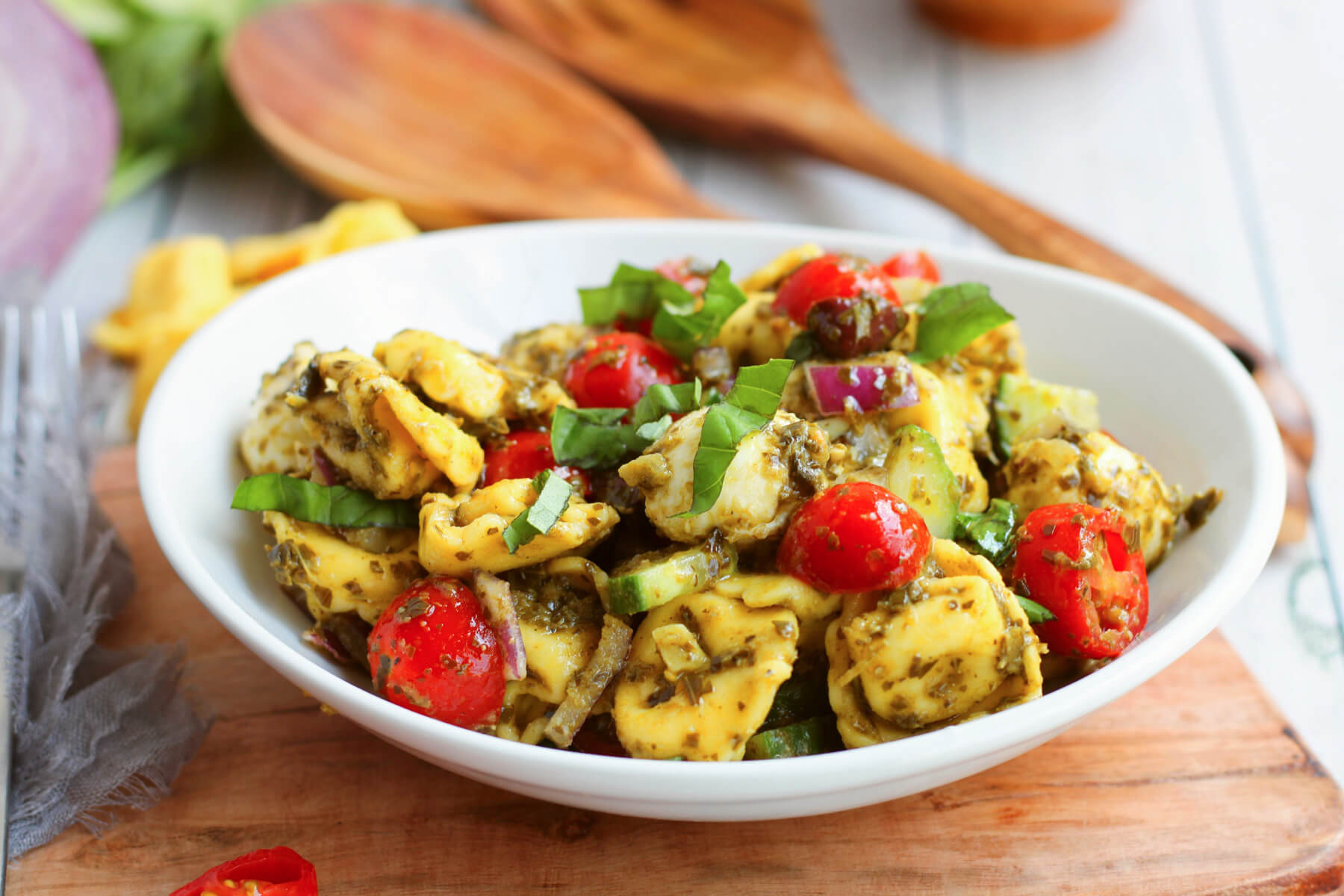 A white bowl filled with brightly coloured Pesto Tortellini Salad below two wooden salad servers