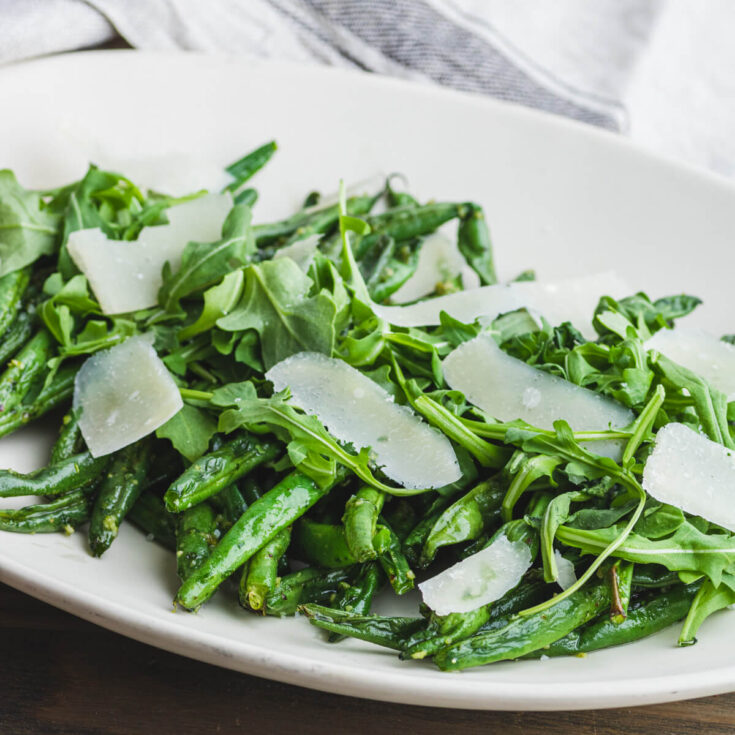 Pesto Green Beans on a white platter topped with baby arugula and Parmesan cheese.