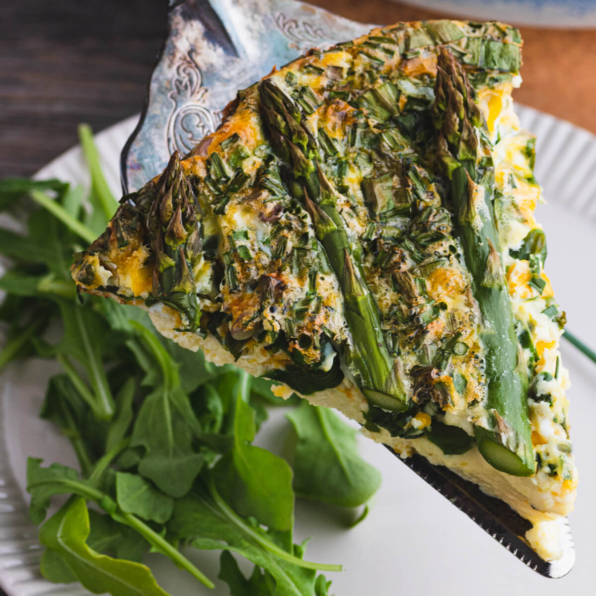 A silver pie server under a slice of Asparagus quiche with wild ramps on a white plate.