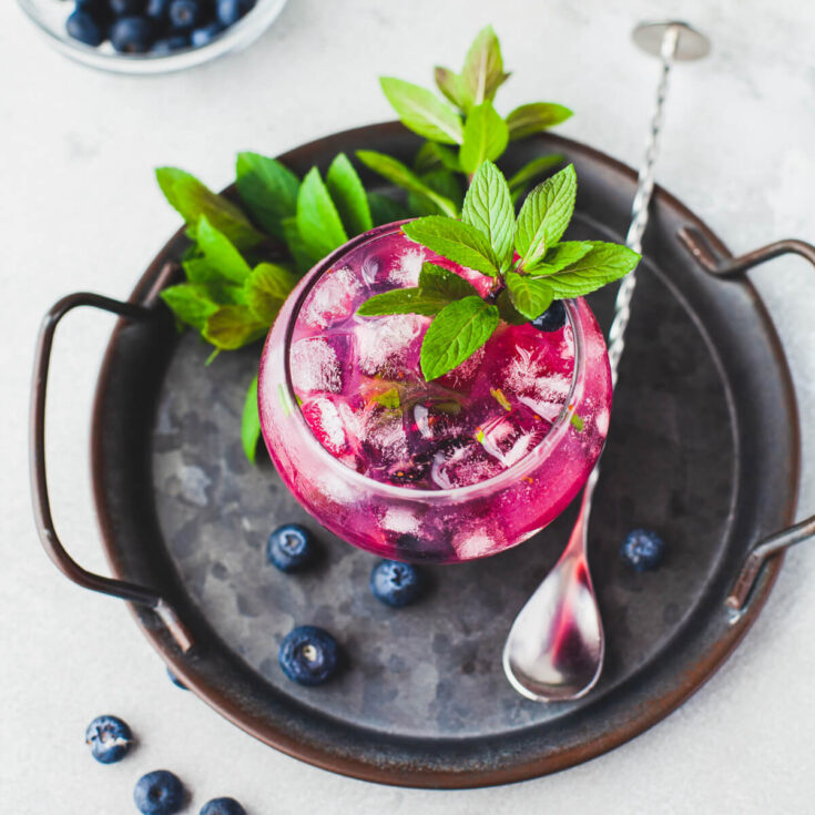 A vibrant purple Blueberry Mojito Cocktail garnished with fresh mint sits on a dark metal tray.