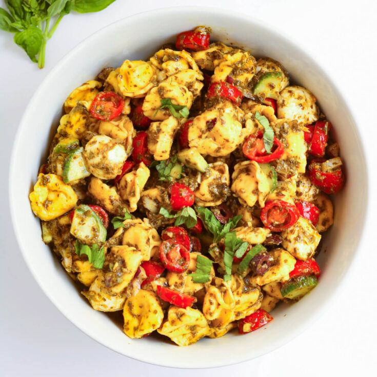 A white bowl filled with tortellini pasta salad - bright vegetables and fresh basil coated in pesto.