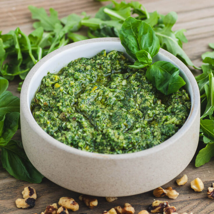 A small stoneware bowl filled with vibrant green arugula pesto on a serving board surrounded by fresh basil, arugula leaves, and walnuts.