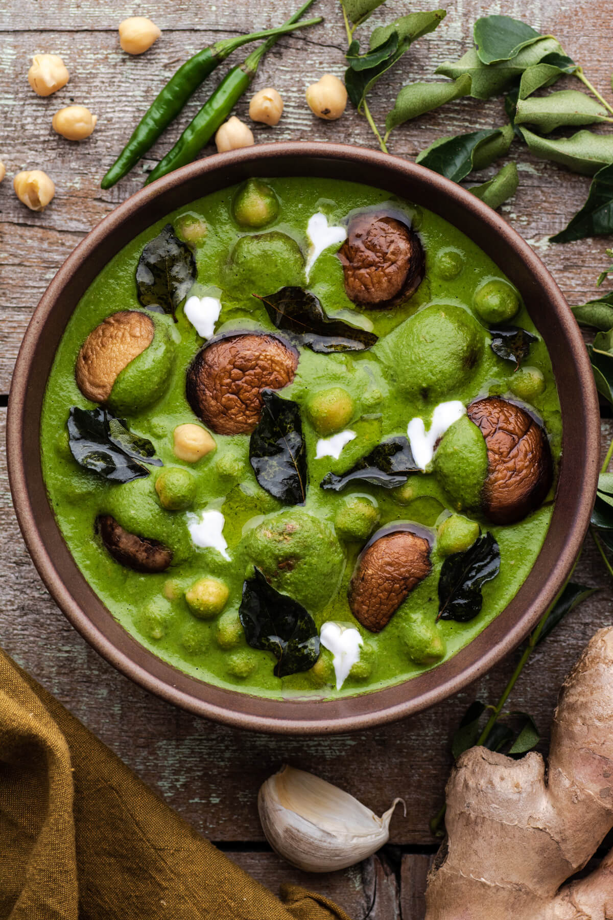 A vibrant green Chana Saag with mushrooms, chickpeas, fried curry leaves, and drizzles of coconut cream in a brown bowl.