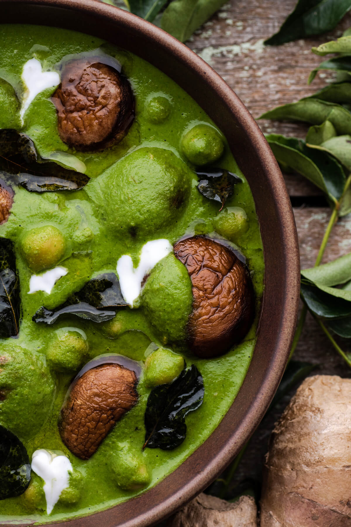 A vibrant green spinach curry with mushrooms, chickpeas, fried curry leaves, and drizzles of coconut cream in a brown bowl.