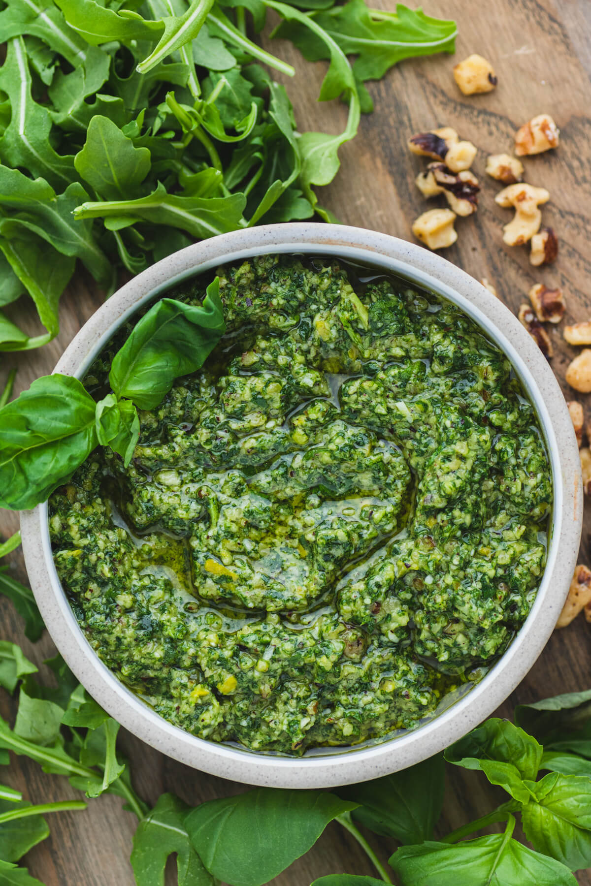 A small stoneware bowl filled with vibrant green arugula pesto on a serving board surrounded by fresh basil, arugula leaves, and walnuts. 