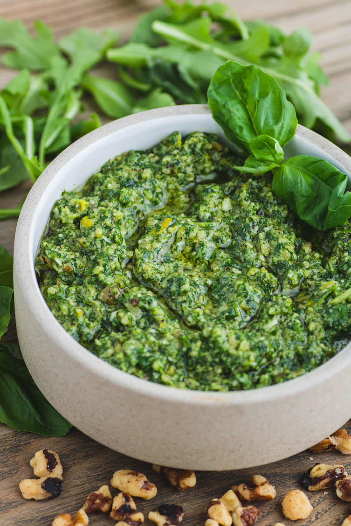 A small stoneware bowl filled with vibrant green arugula pesto on a serving board surrounded by fresh basil, arugula leaves, and walnuts.