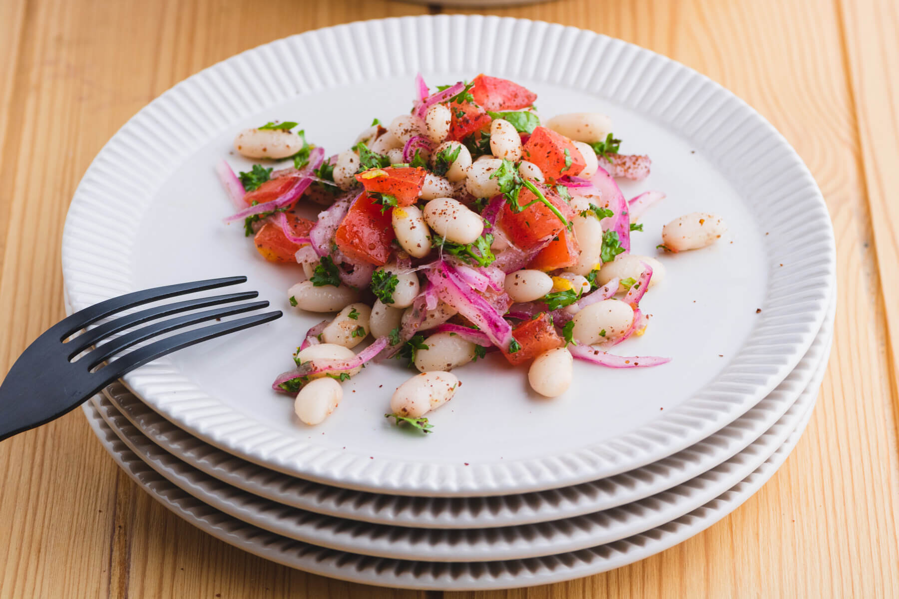 A serving of colourful white bean salad with red onions, tomatoes, and green curly parsley on a white plate.